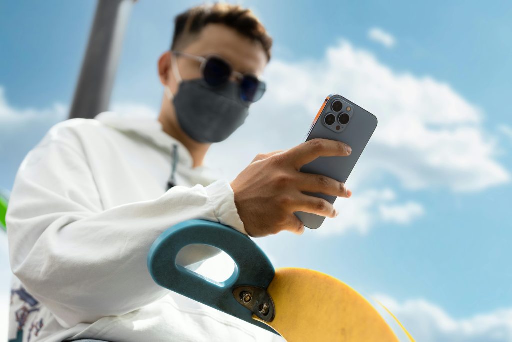 A young man wearing a face mask uses his cellphone outdoors with a skateboard in the foreground.